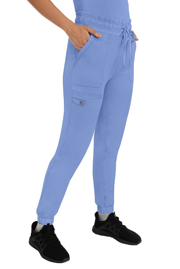 A female LPN wearing an HH Works Women's Renee Jogger Scrub Pant in Ceil featuring 2 front patch pockets & 1 cargo pocket the wearer's right side leg.