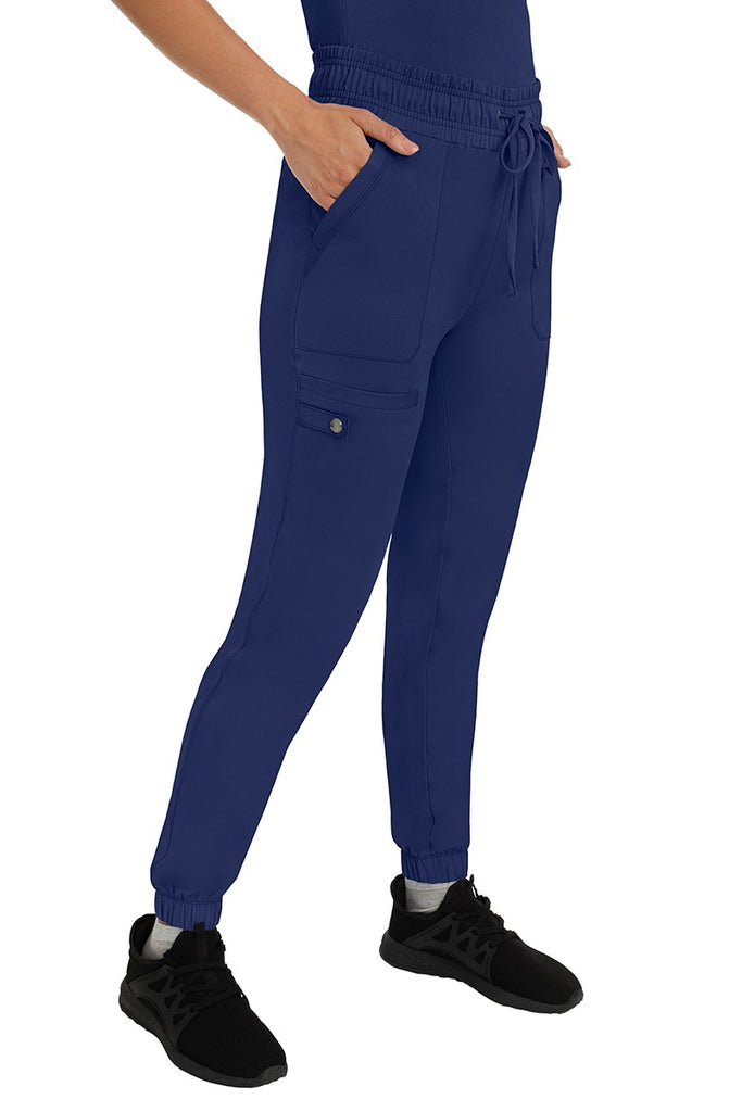A female LPN wearing an HH Works Women's Renee Jogger Scrub Pant in Navy featuring 2 front patch pockets & 1 cargo pocket the wearer's right side leg.