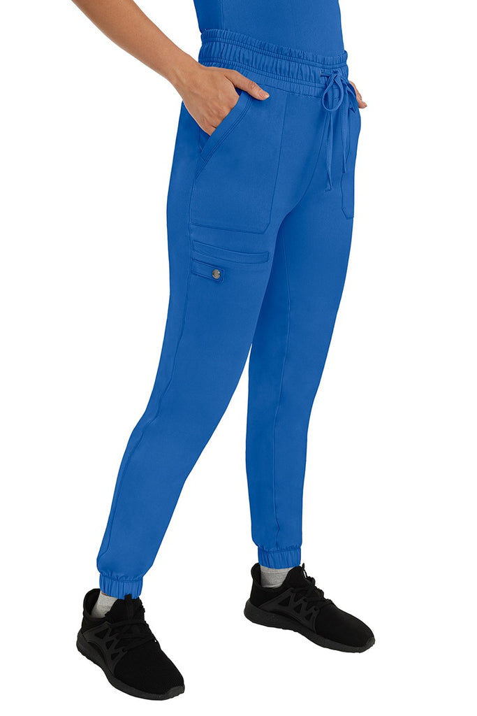 A female LPN wearing an HH Works Women's Renee Jogger Scrub Pant in Royal featuring 2 front patch pockets & 1 cargo pocket the wearer's right side leg.