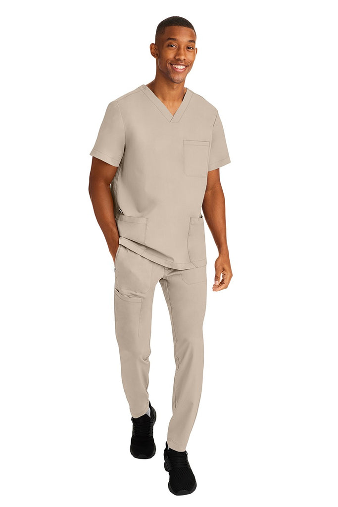 A young male RN wearing an HH-Works Men's Ryan Multi-Pocket Cargo Scrub Pant in Khaki featuring an easy care fabric that is quick drying, moisture wicking, and ultra-flexible.